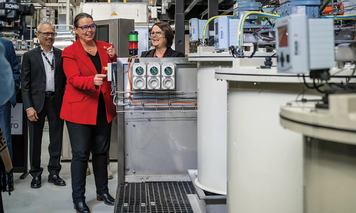 A woman in a red jacket is surrounded by tanks and other industrial equipment in a chemistry laboratory.