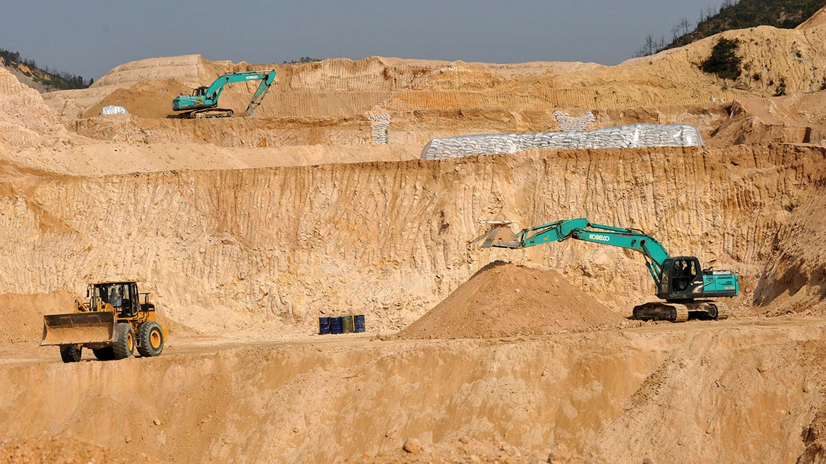Two turquoise-colored excavators and a yellow bulldozer move yellow-brown sediment in a rare earth mining pit with several levels.