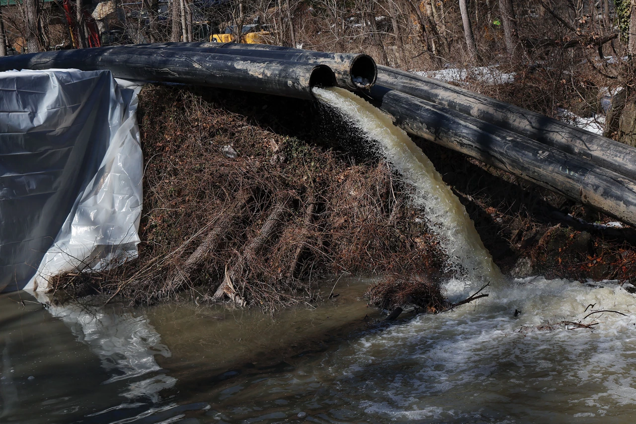 Water spills out of an open sewer pipe into a body of water in a wooded area, with another open sewer pipe beside the first.
