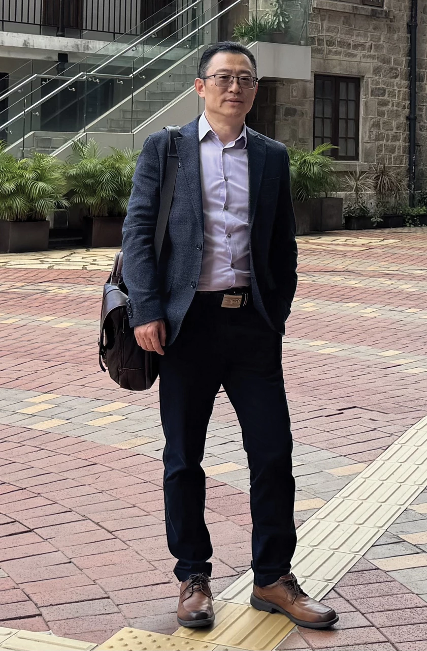 A person in a blazer and dress shirt stands on paving stones in front of a building.