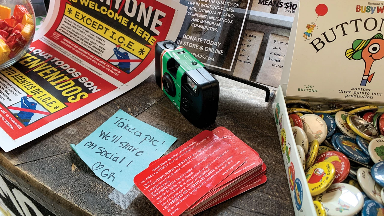 A green-and-black disposable camera sits on a retail counter next to a small sign that reads, “Take a pic! We’ll share on social! ♡ GR”
