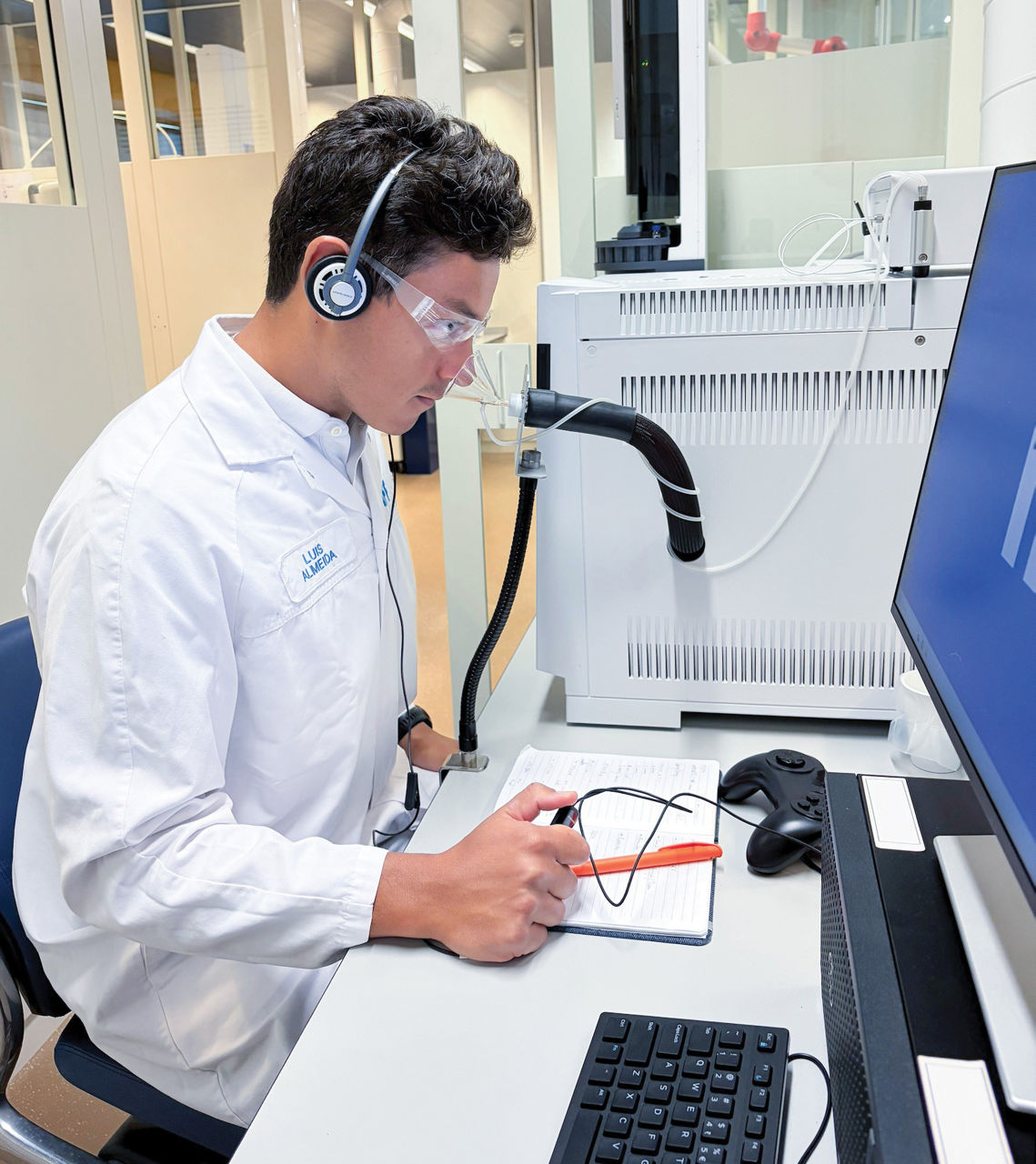 A person sitting at a desk in a white lab coat, safety goggles, and headphones sticks their nose into a clear, plastic cone attached to a box-shaped instrument while holding a black remote.