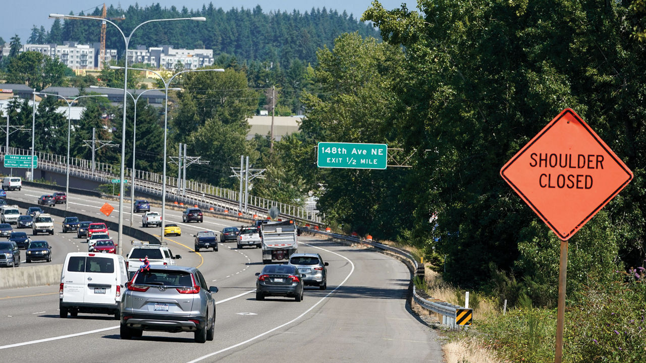 Cars winding down a Washington highway with trees and buildings in the background. There is an orange “shoulder closed” sign as well as a green sign for a nearby exit.