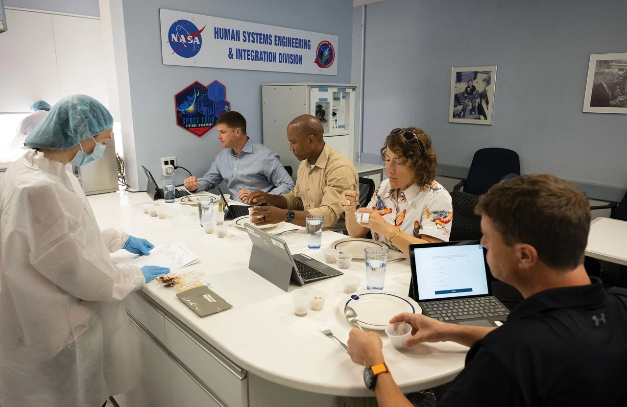 Four people sit around a table, eating food from small cups or looking at their laptop screens, while another person wearing a lab coat, gloves, and mask stands on the other side of the table looking at a sheet of paper. The table has several cups, plates, and packets of food.