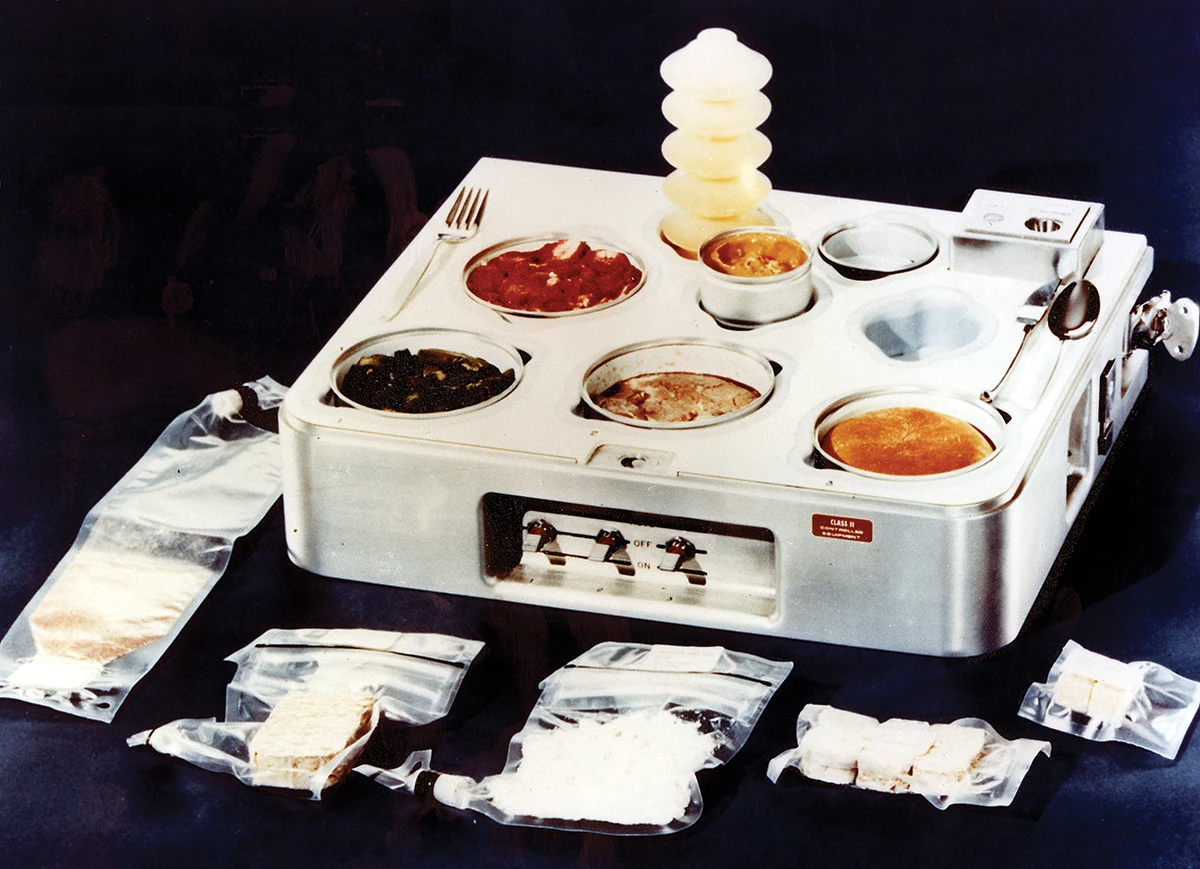 A heating tray for an early NASA space station shows multiple bowls of different foods of varying colors. A number of packages of dehydrated foods in plastic bags are lying around the tray.