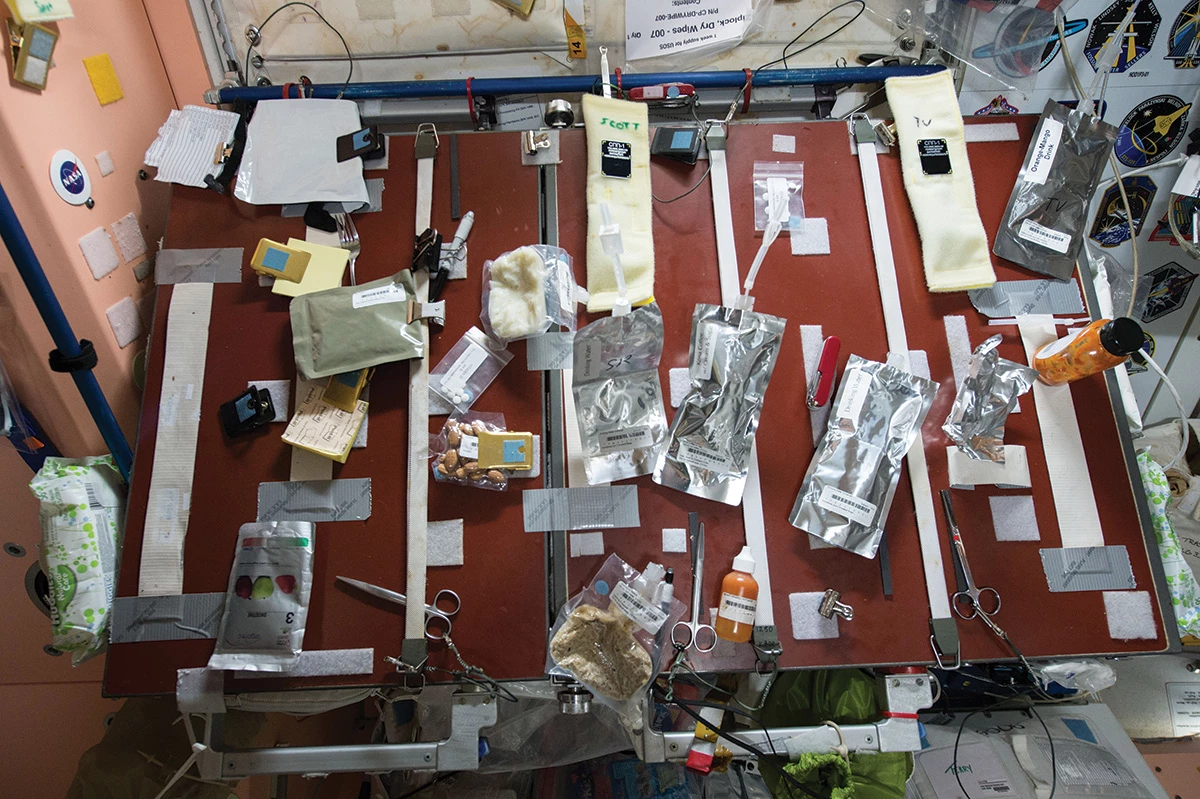 Several packets of food and drink items in silver packaging attached to a table with clips and tape.