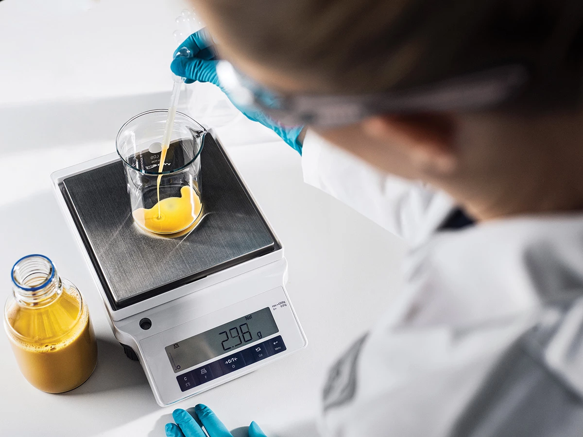 Top view of a person wearing gloves and safety goggles uses a dropper to pour a yellow liquid into a beaker on a scale.