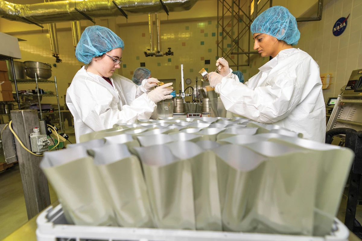 Several open pouches of food are in the forefront, while two workers wearing lab coats, caps, and gloves fill other pouches with food in the background.
