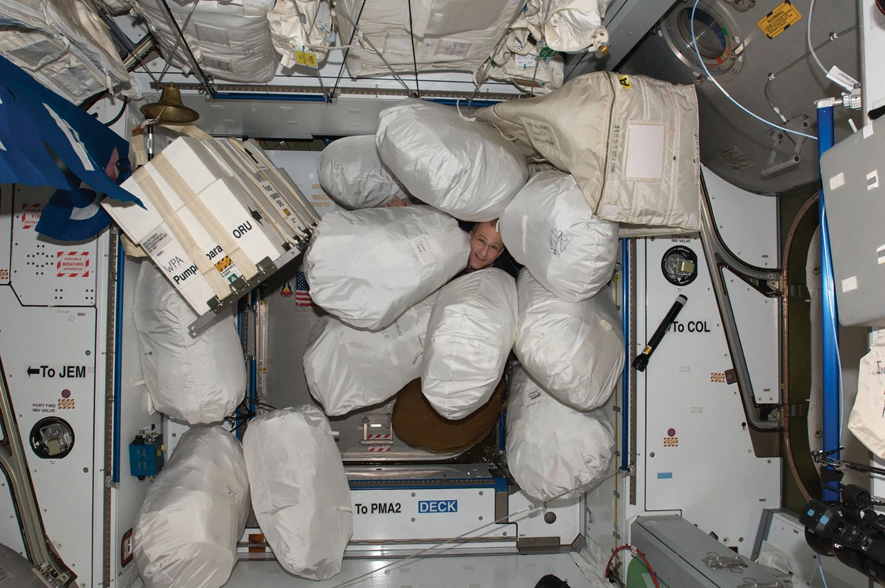 An astronaut’s face looks out through a gap between several large, stuffed bags that are floating without gravity.