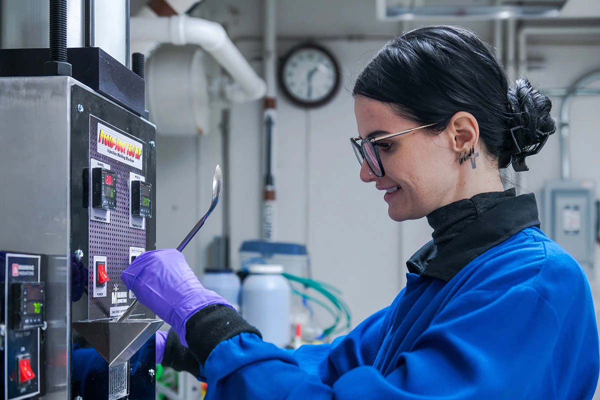 A scientist in a blue lab coat, glasses, and purple gloves stands in front of a machine with digital numbers on it and holds a long, thin tool against the machine, with a clock and some pipes in the background.
