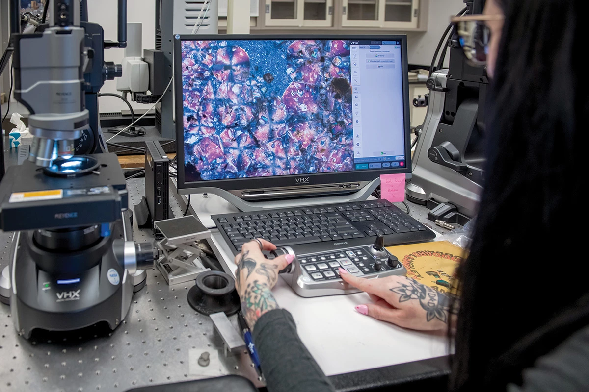 A scientist sits at a computer, turning a knob and pushing a button on a small device in front of the computer, while the computer screen shows an image with blotches of red and blue. To the left on the table is a microscope with a light illuminating a sample under the lens.