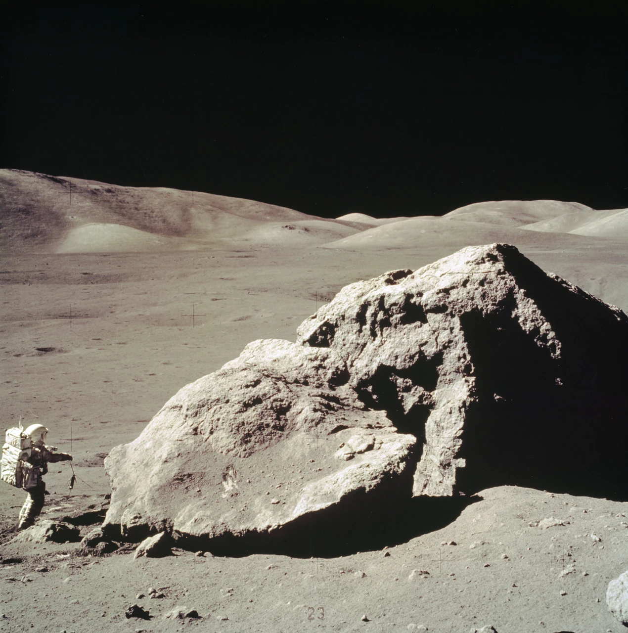 A lunar astronaut stands to the left of a large rock. The sky is black and the land is gray.