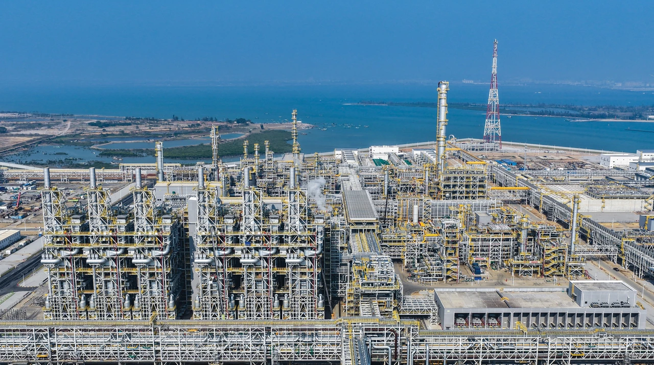A large petrochemical plant with gray scaffolding and yellow piping stands near a coastline.