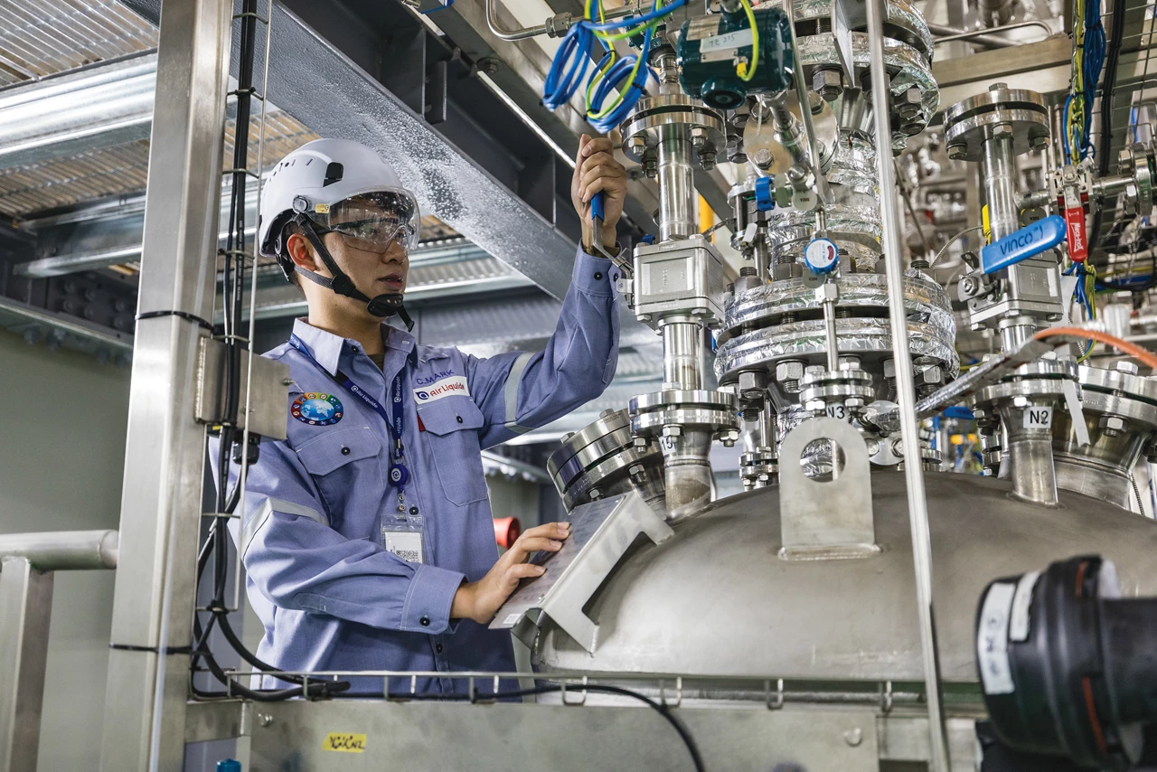 A person in a hard hat and uniform handles a lever on a small chemical reactor.