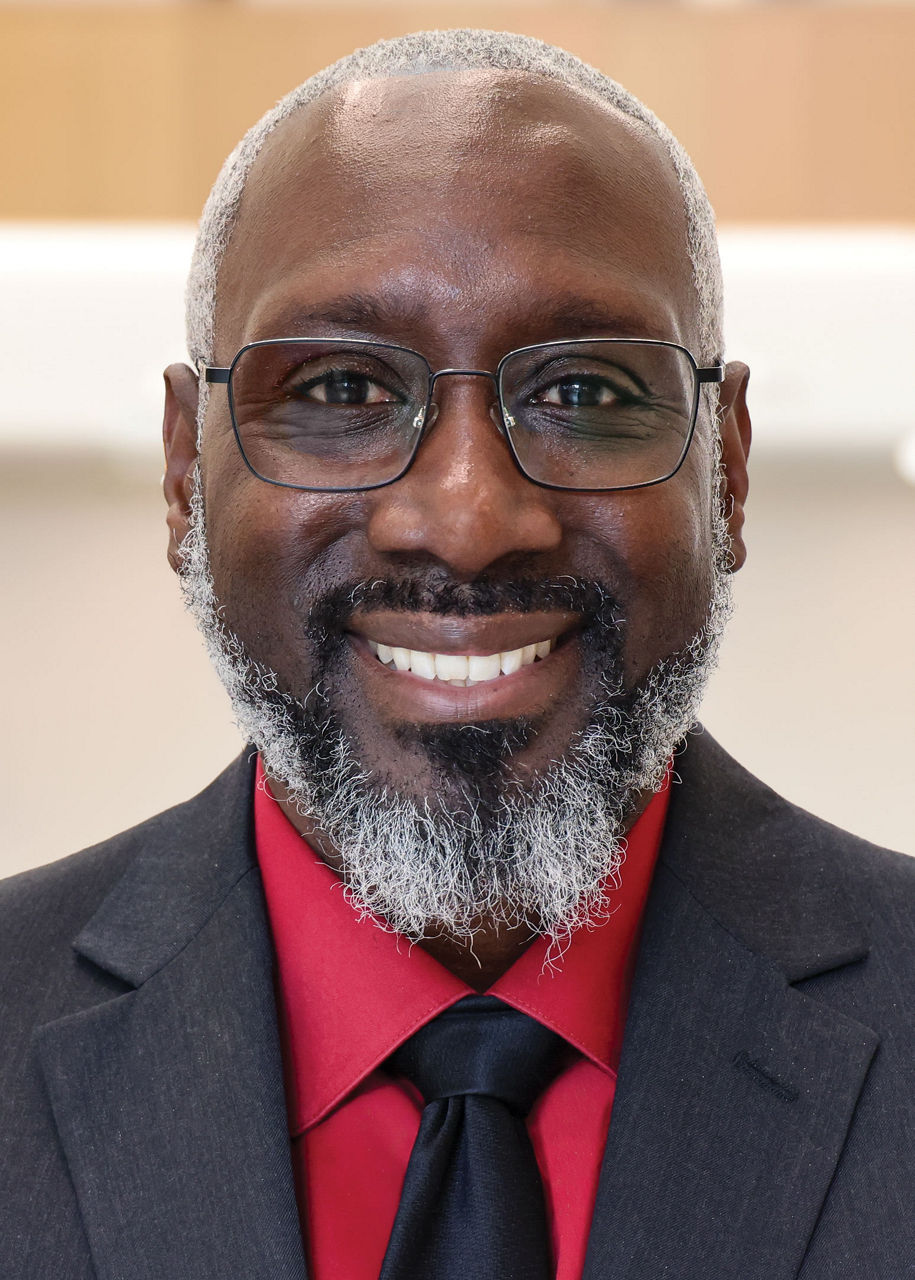 A man wearing glasses, a red dress shirt, and a dark tie and suit jacket smiles for a portrait shot.