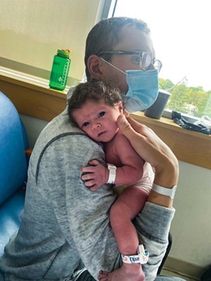 Um homem vestindo óculos e uma máscara cirúrgica segura um bebê recém -nascido com cabelos escuros encaracolados e grandes olhos castanhos e usando uma pulseira de hospital. A configuração parece ser uma sala em uma suíte de maternidade.