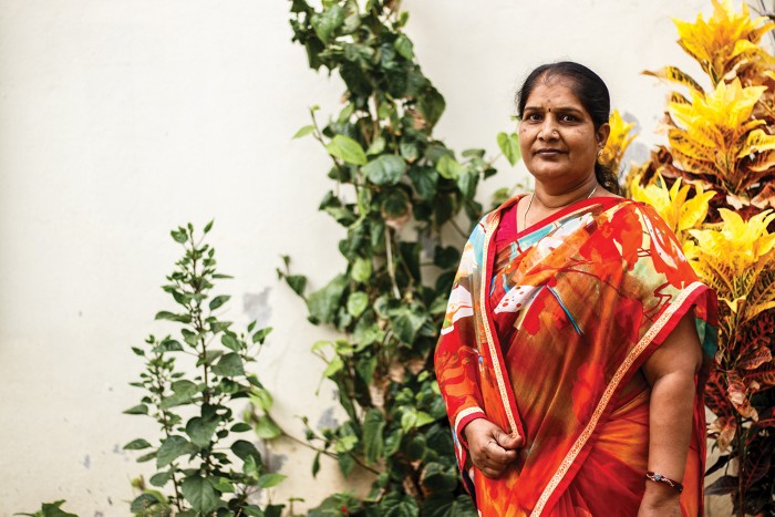 Sunita Bhosale wears and red and orange sari and stands in front of tall plants growing against a light-colored wall.
