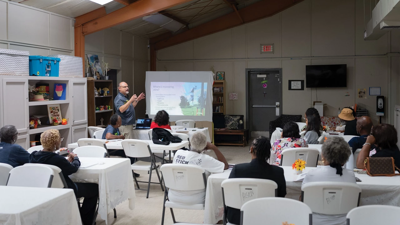 A man wearing a blue shirt delivers a presentation in front of seated participants.