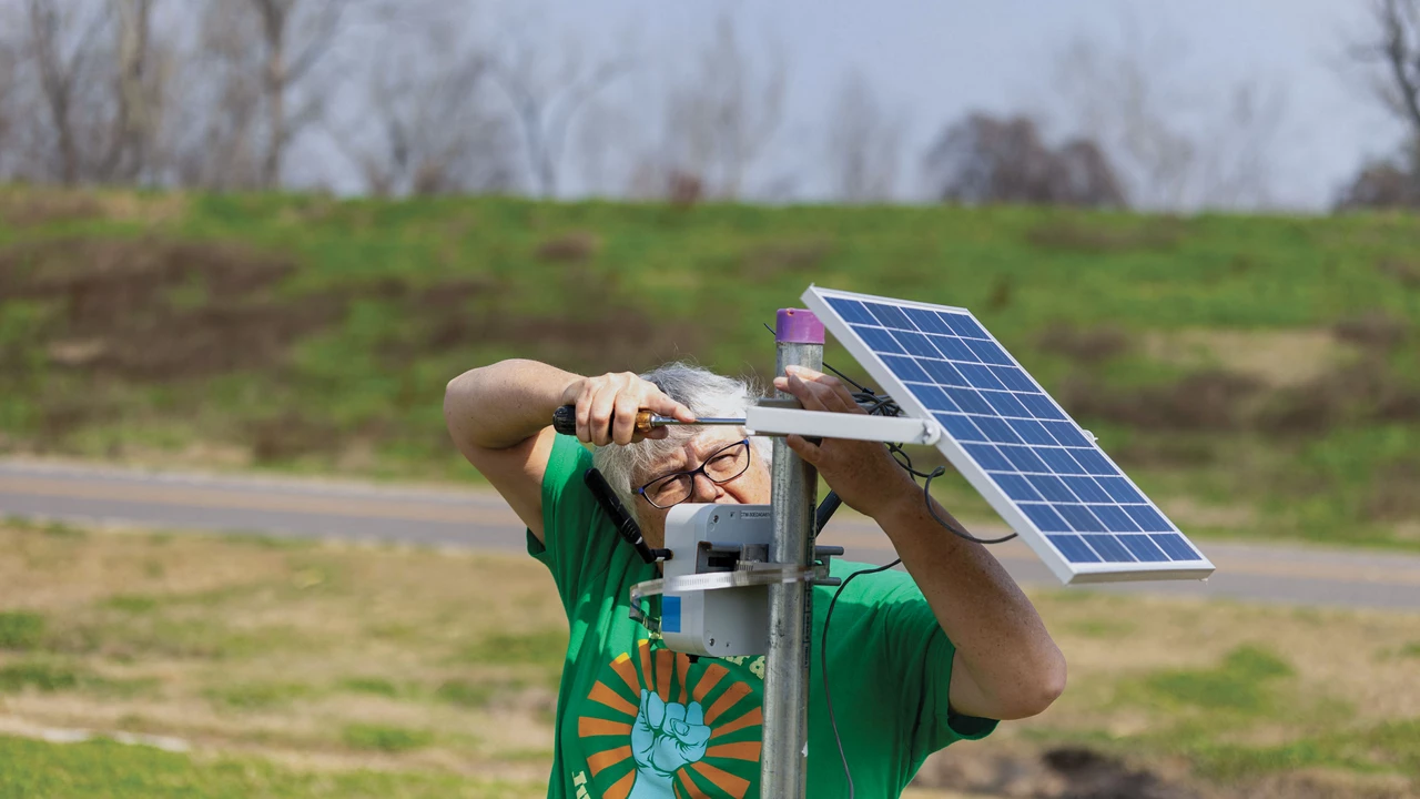 Woman with short gray hair, wearing glasses and a green T-shirt, uses a screwdriver to work on a blue panel attached to a metal pole.