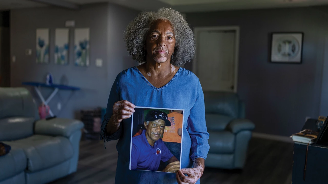 Woman wearing a blue shirt and standing in a living room holds a framed photo of a man.