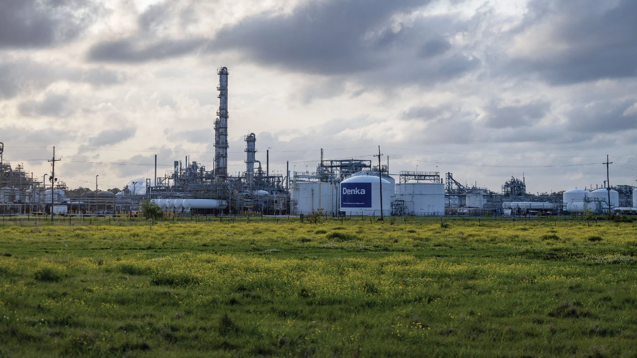 An industrial facility with a smokestack and a group of low white buildings behind a field of grass.