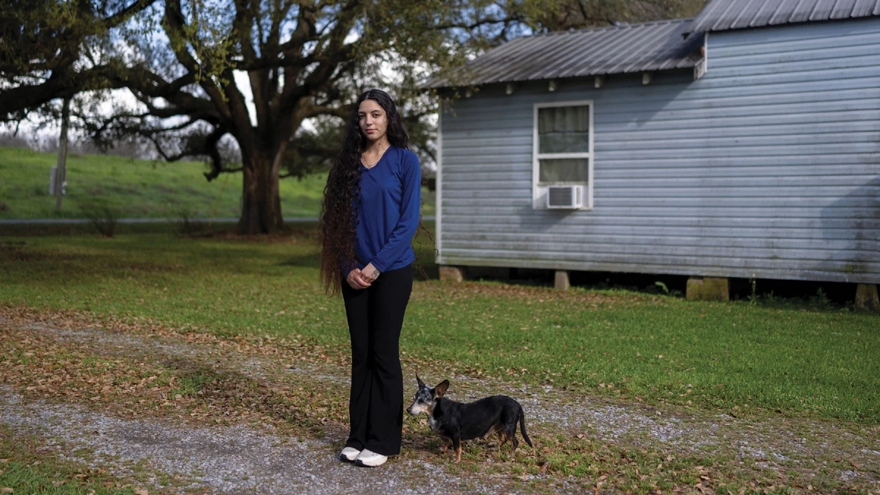 Woman wearing dark trousers and a blue shirt stands with a small dog, with a gray house and a tree in the background.