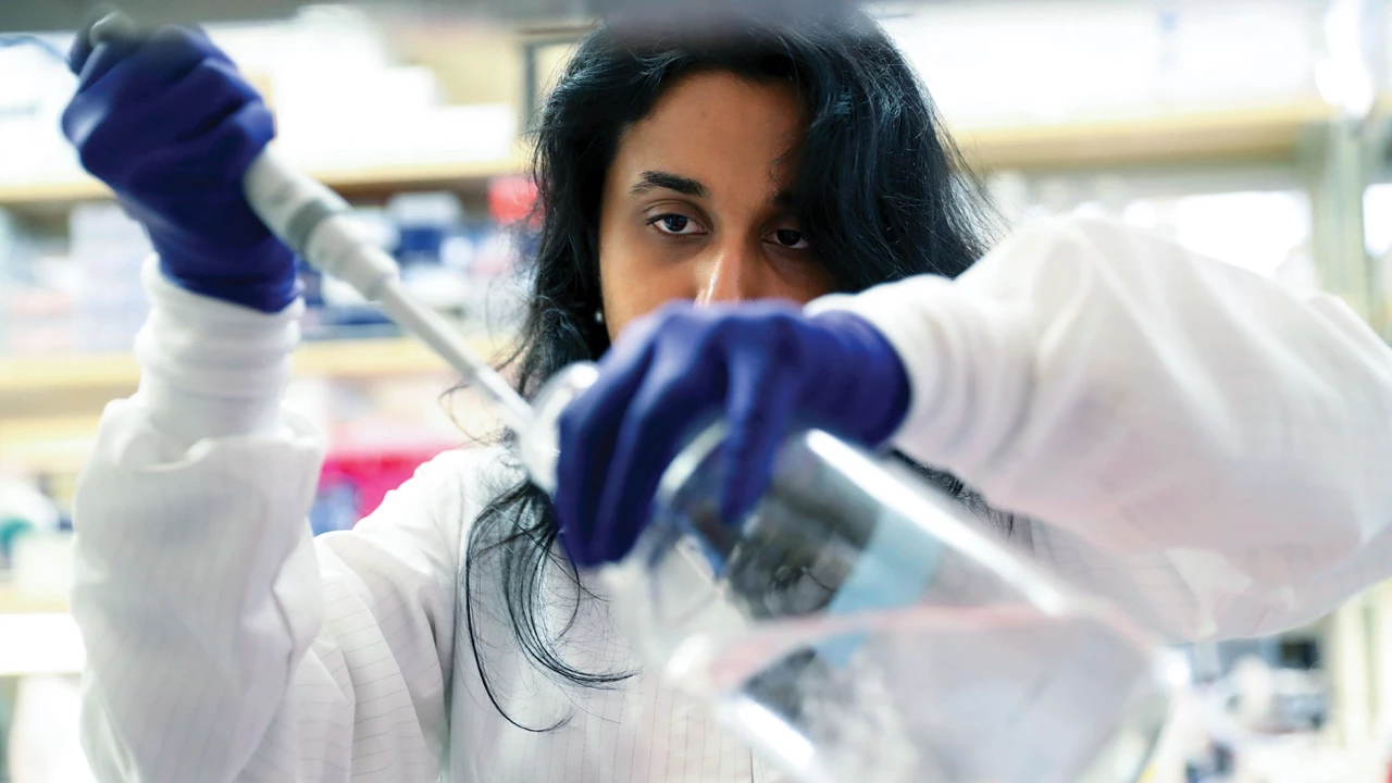 A woman wearing a lab coat and blue safety gloves injects a substance into a bottle.
