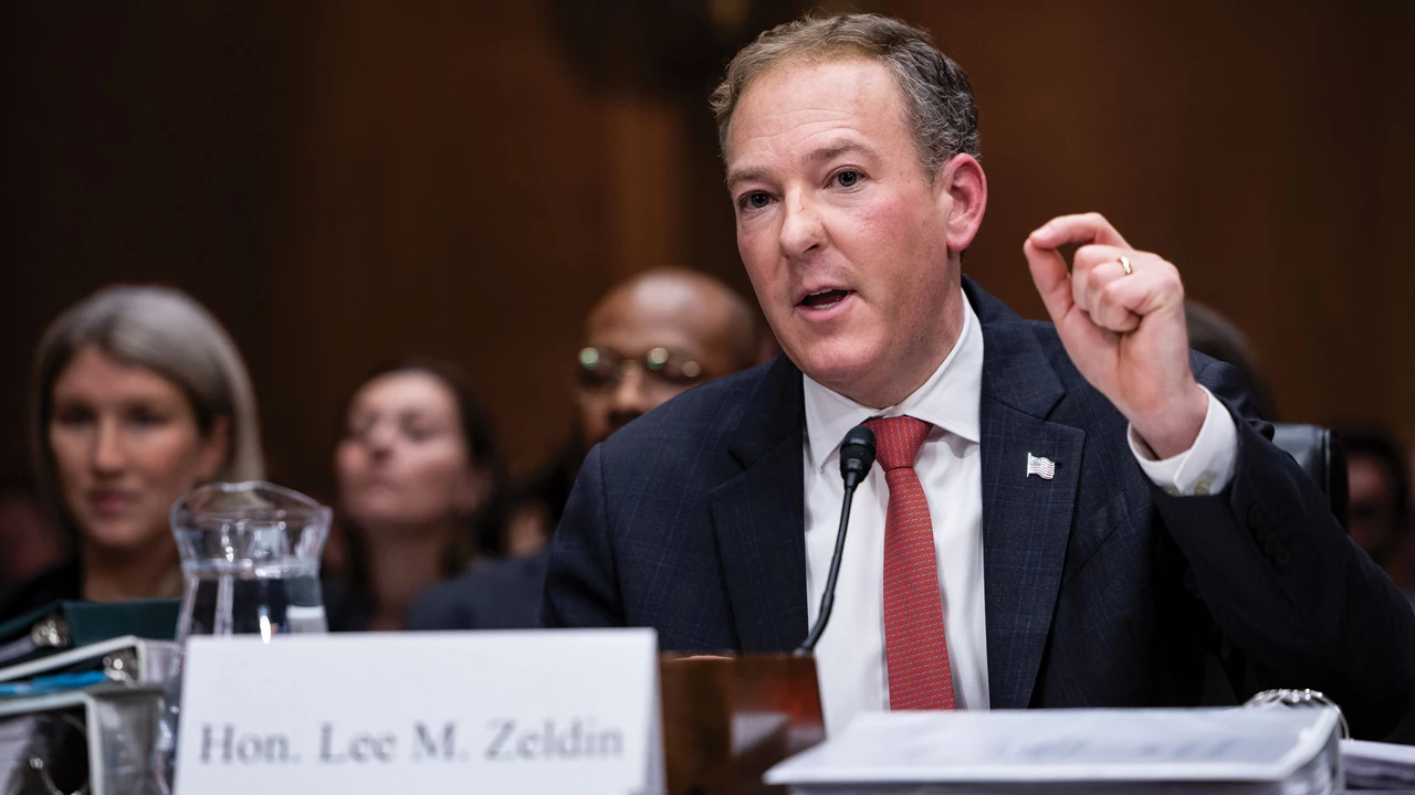 Lee Zeldin wears a suit and tie and sits at a table in front of a microphone, and several people sit behind him.