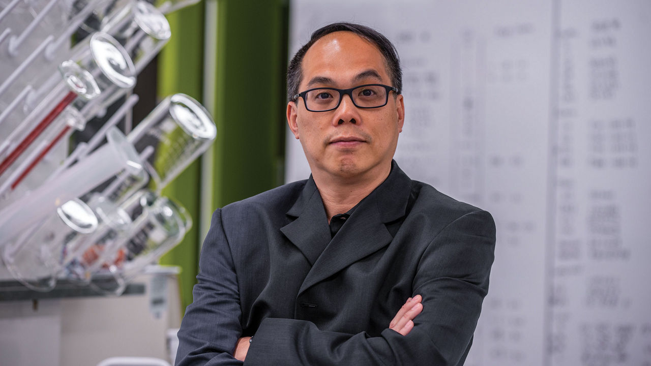A man wearing glasses and a dark jacket stands with crossed arms in a lab with a white board in the background and tubes to the left foreground.