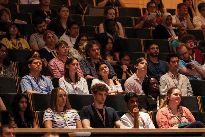 Várias dezenas de pessoas, muitas delas jovens e todas vestindo crachás de participantes, sentam -se de frente para a frente nos assentos do auditório.