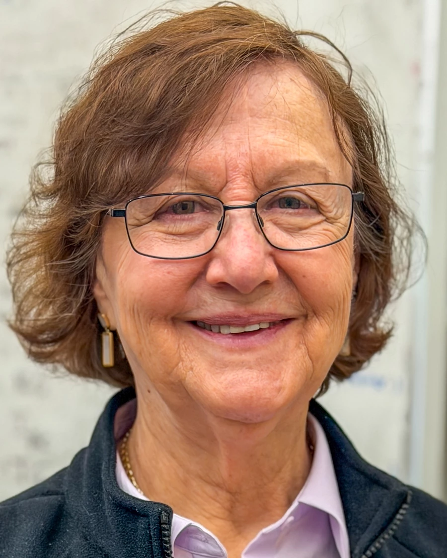 A woman in a purple collared shirt, sleeveless gray fleece vest, and glasses smiles at the camera.