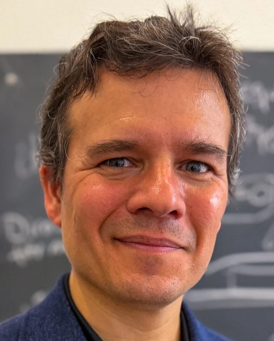 A man in a black collared shirt and dark-blue blazer smiles at the camera.