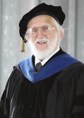 Dean F. Martin wears a black graduation gown with blue hood, a black floppy hat with a gold tassel, and glasses. 