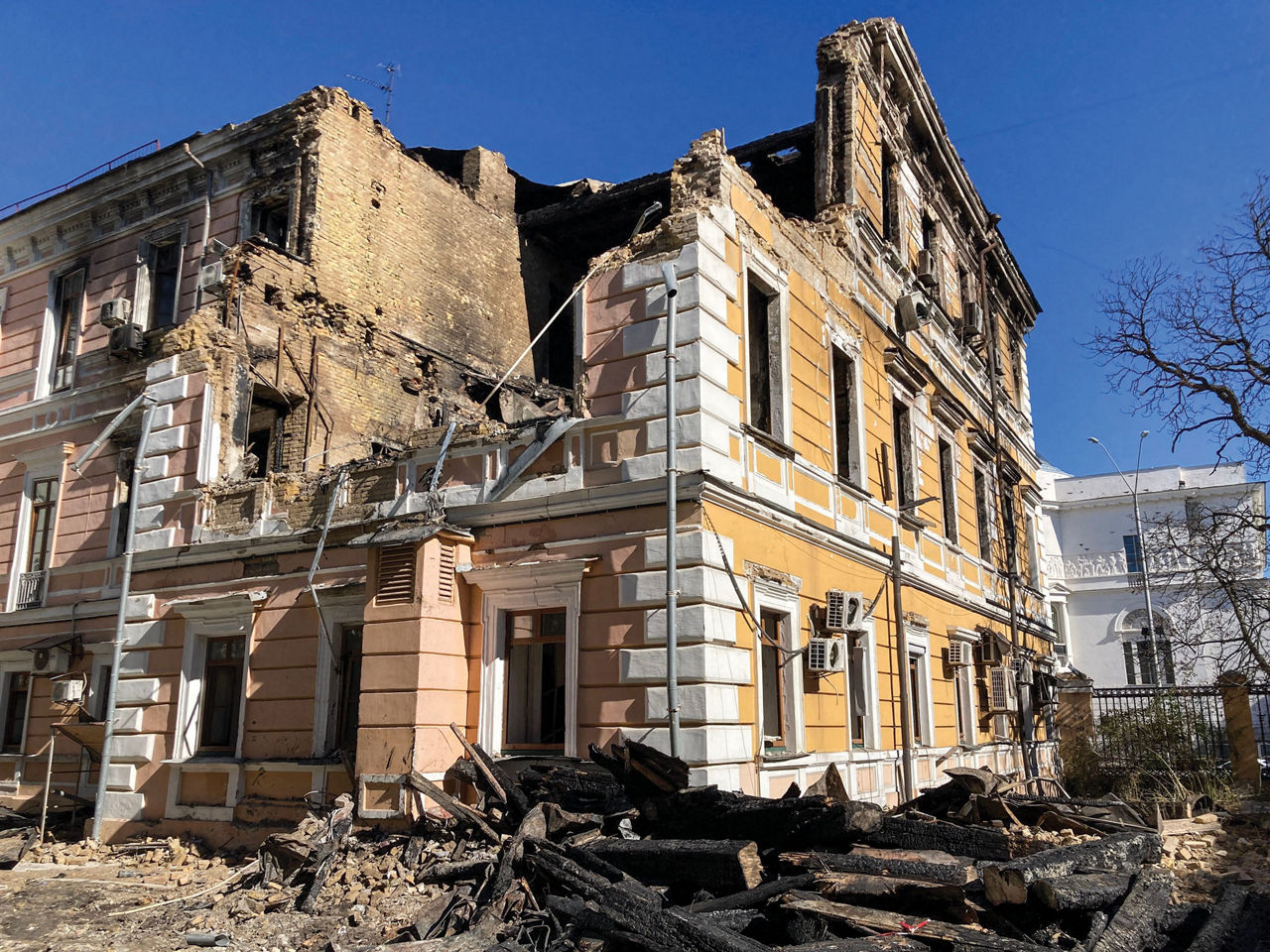 A three-story white, yellow, and salmon-colored building shows significant damage, with parts of the exterior missing and debris lying on the surrounding land.