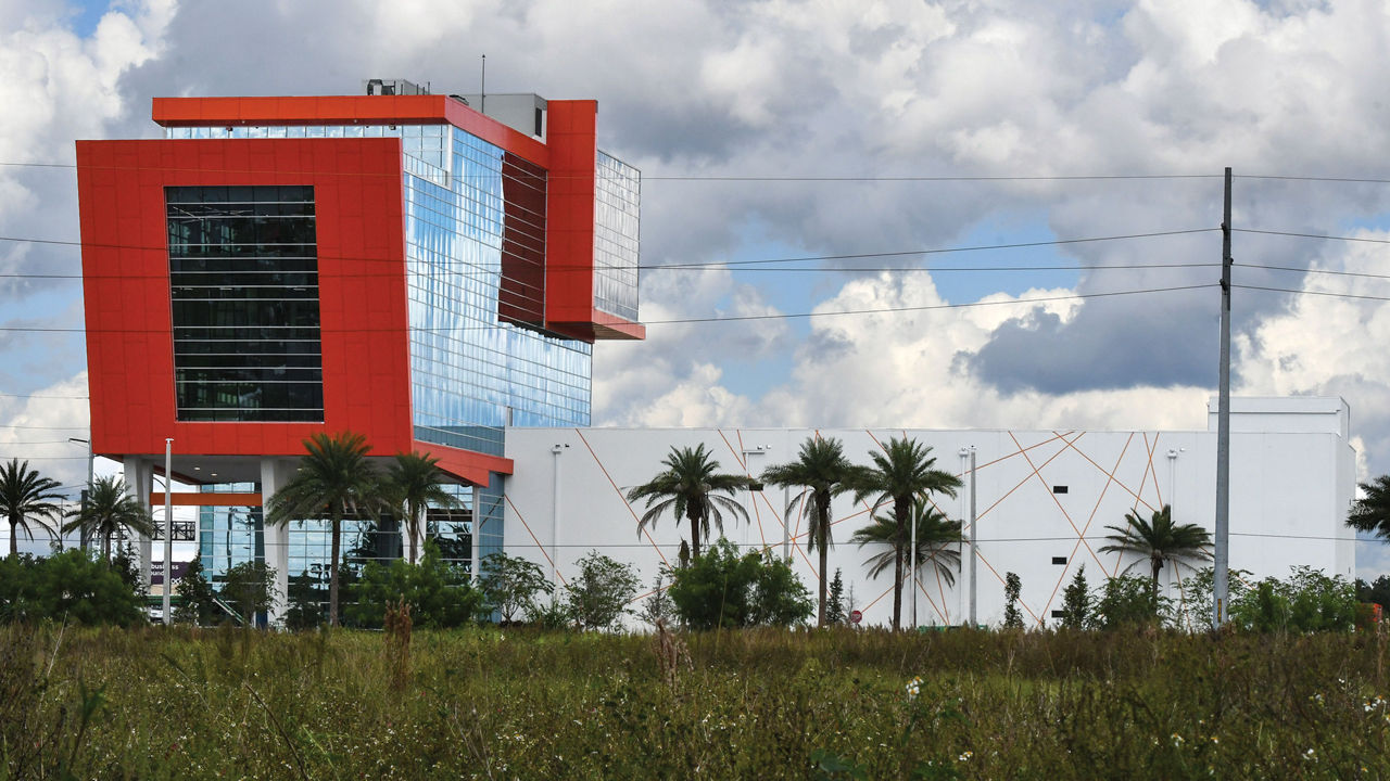 A white rectangular building sits next to a larger glass building with red-orange details. Several palm trees are in the foreground.