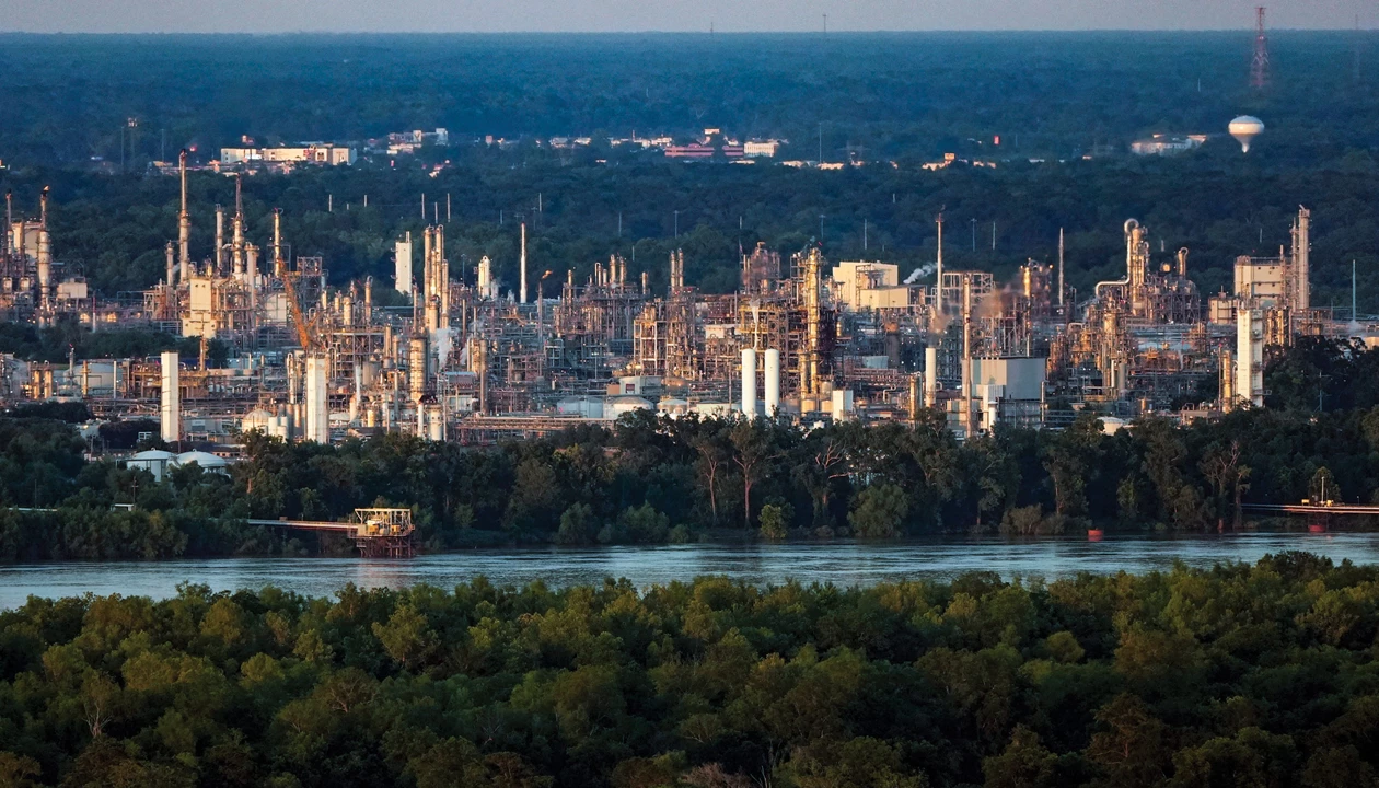 A wide external shot of chemical and other industrial facilities, with a body of water and trees in the foreground.