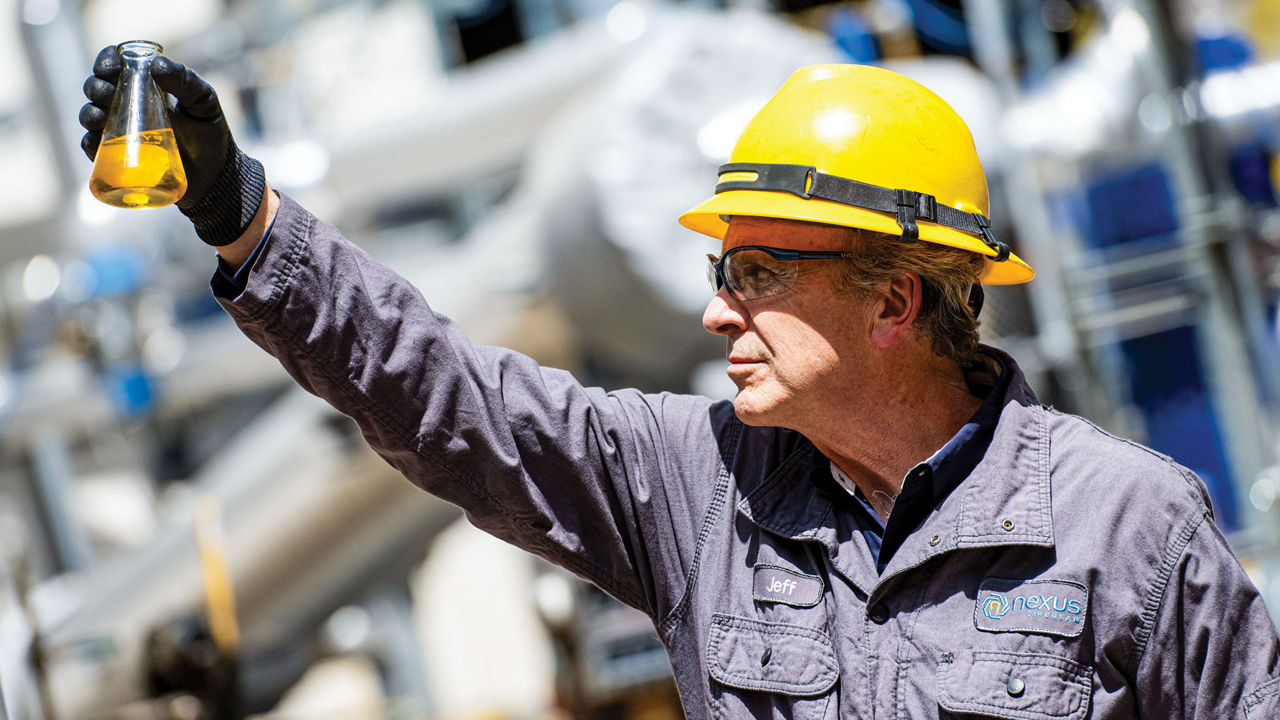 A man in a blue work suit examines a vial of liquid. His nametag on the left says “Jeff,” and the company name, Nexus Circular, is in blue lettering on the right.