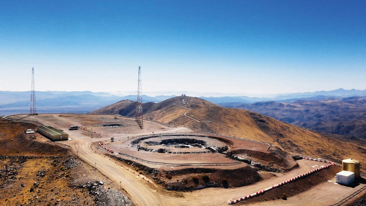 An aerial view of a construction site near a mountain peak. More mountains are visible in the background.