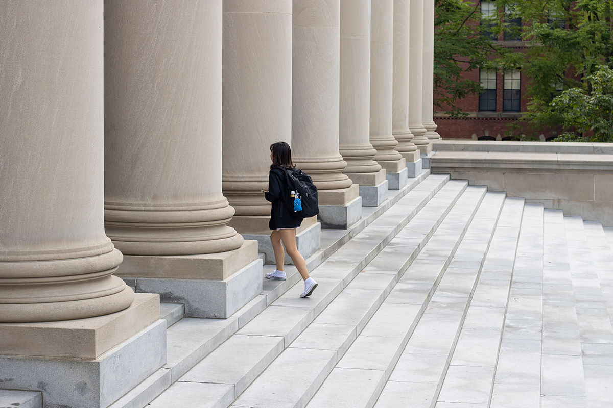 A person, wearing a jacket and carrying a backpack, walks up the steps toward a building with several large exterior columns.