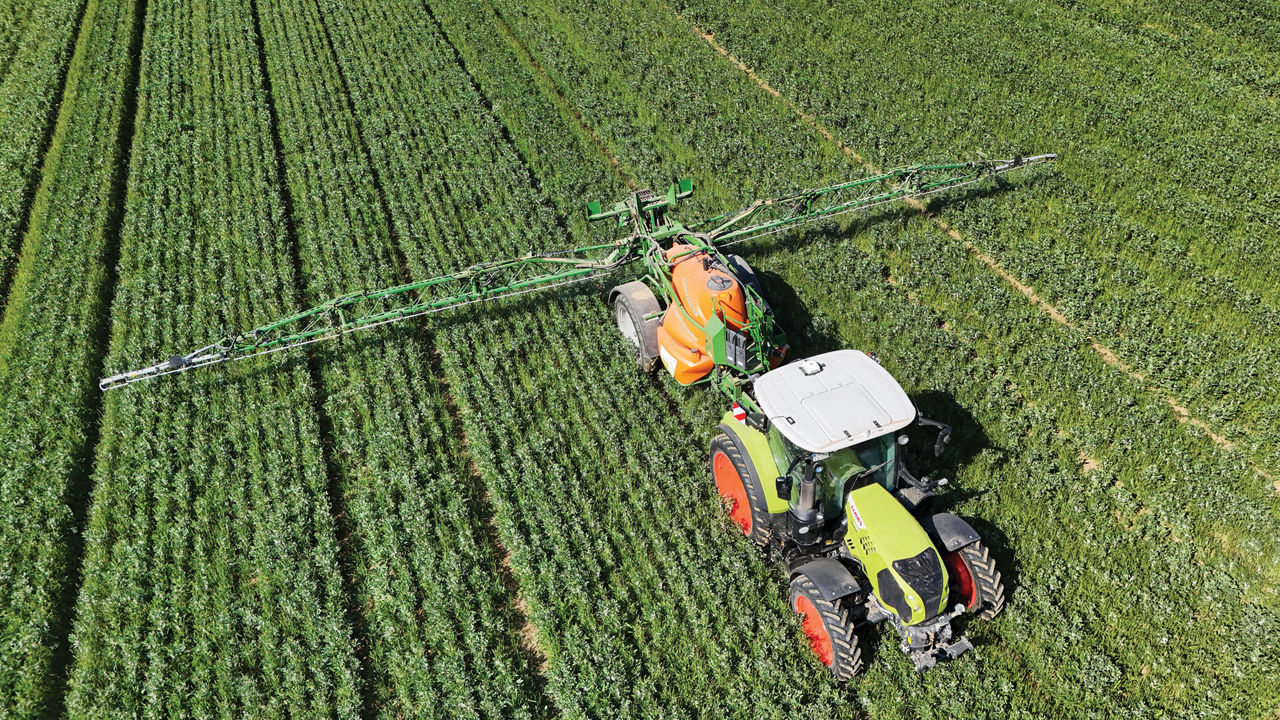 A green-and-orange farm tractor pulls spraying equipment across a green field.