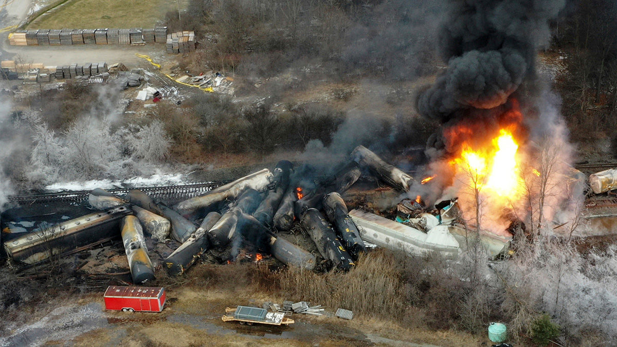 Multiple severely damaged train cars lie in a field, with some of the cars burning and black smoke rising into the air.