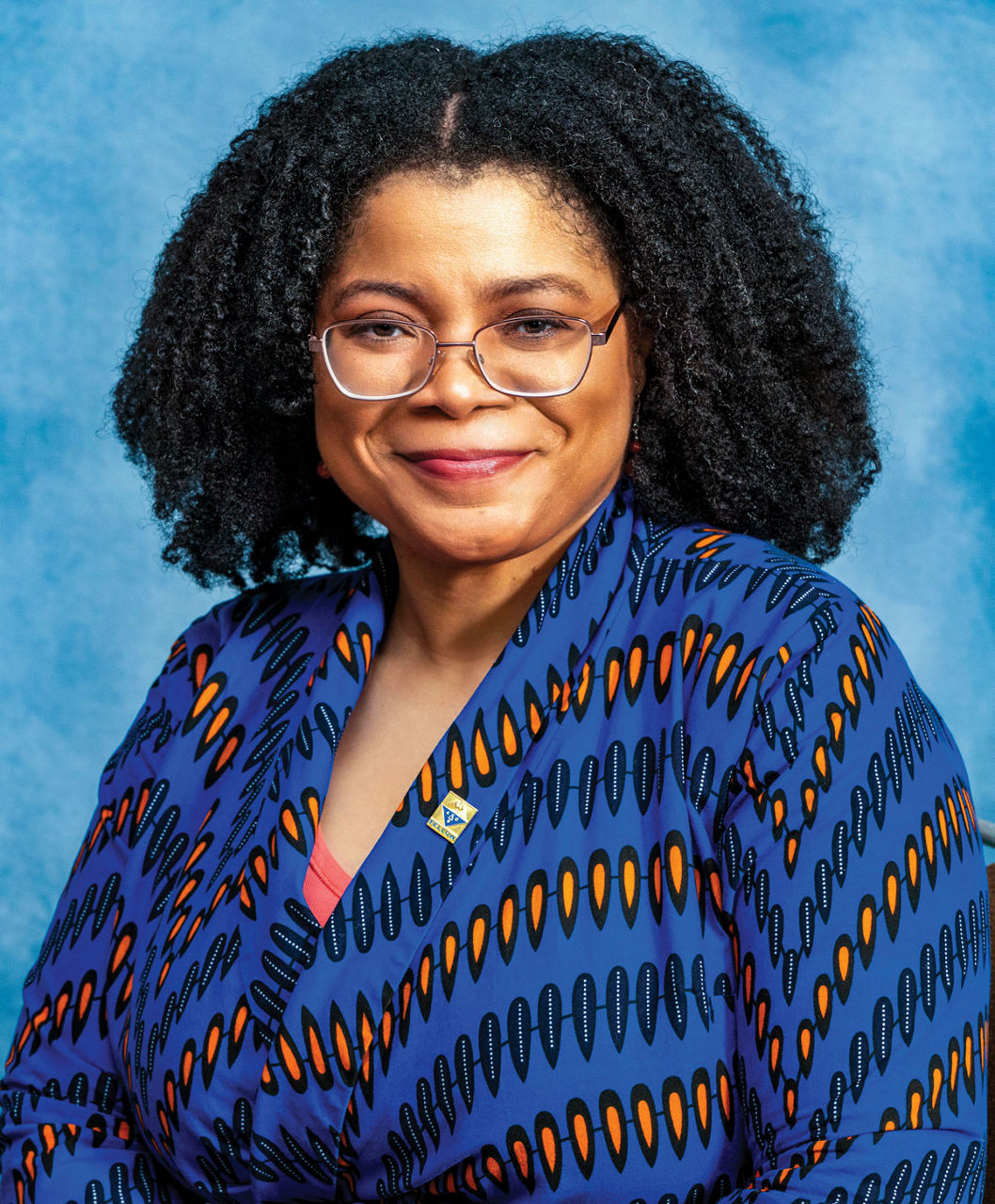 Ann wears a blue, black, and yellow patterned shirt, glasses, and smiles at the camera.