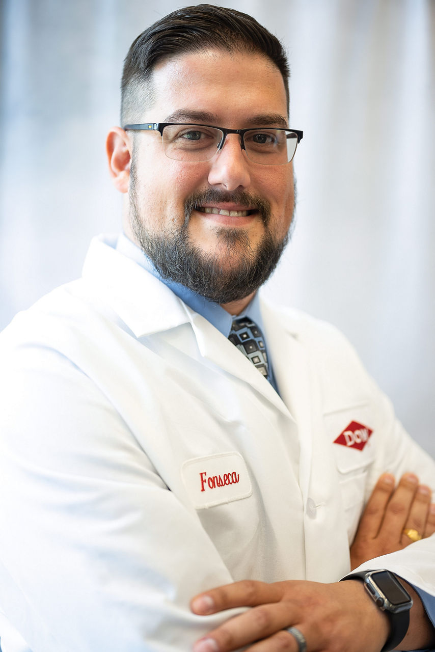 Daniel wears a Dow lab coat, a light-blue collared shirt, a tie, and dark glasses and smiles at the camera for a portrait.