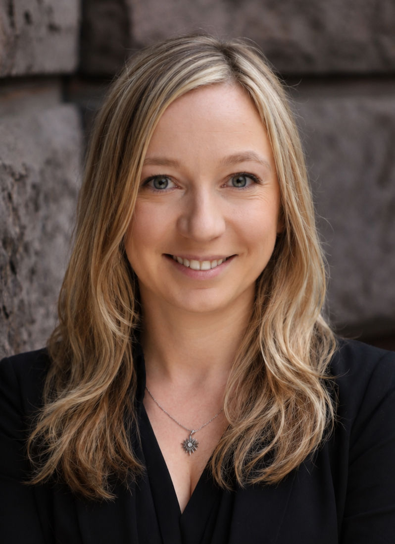Ivona wears a black suit jacket, and with her arms crossed smiles at the camera.