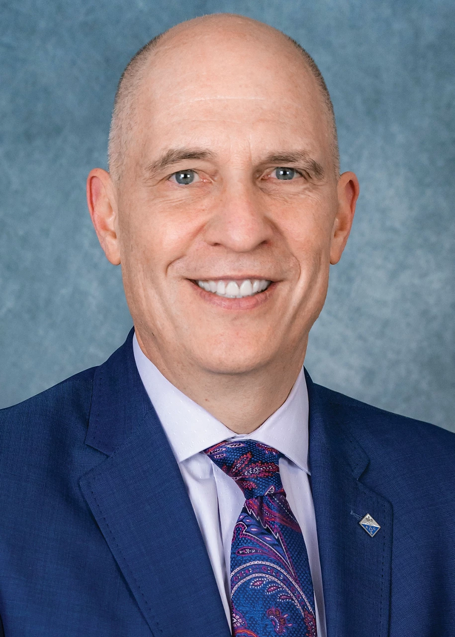 A man wears a blue blazer and blue paisley tie and smiles at the camera.