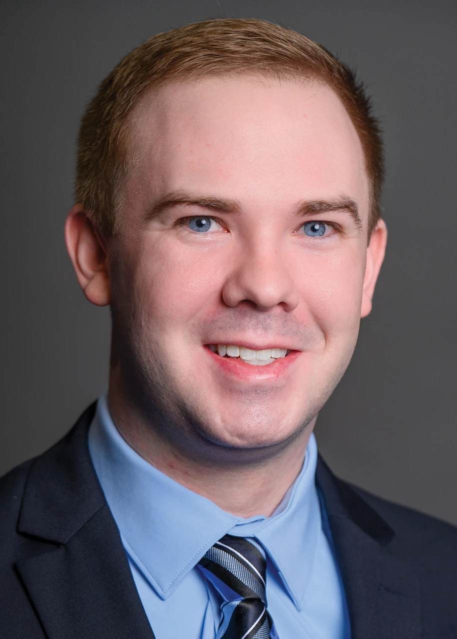 Sam wears a blazer, light blue shirt, and striped blue tie and smiles at the camera.