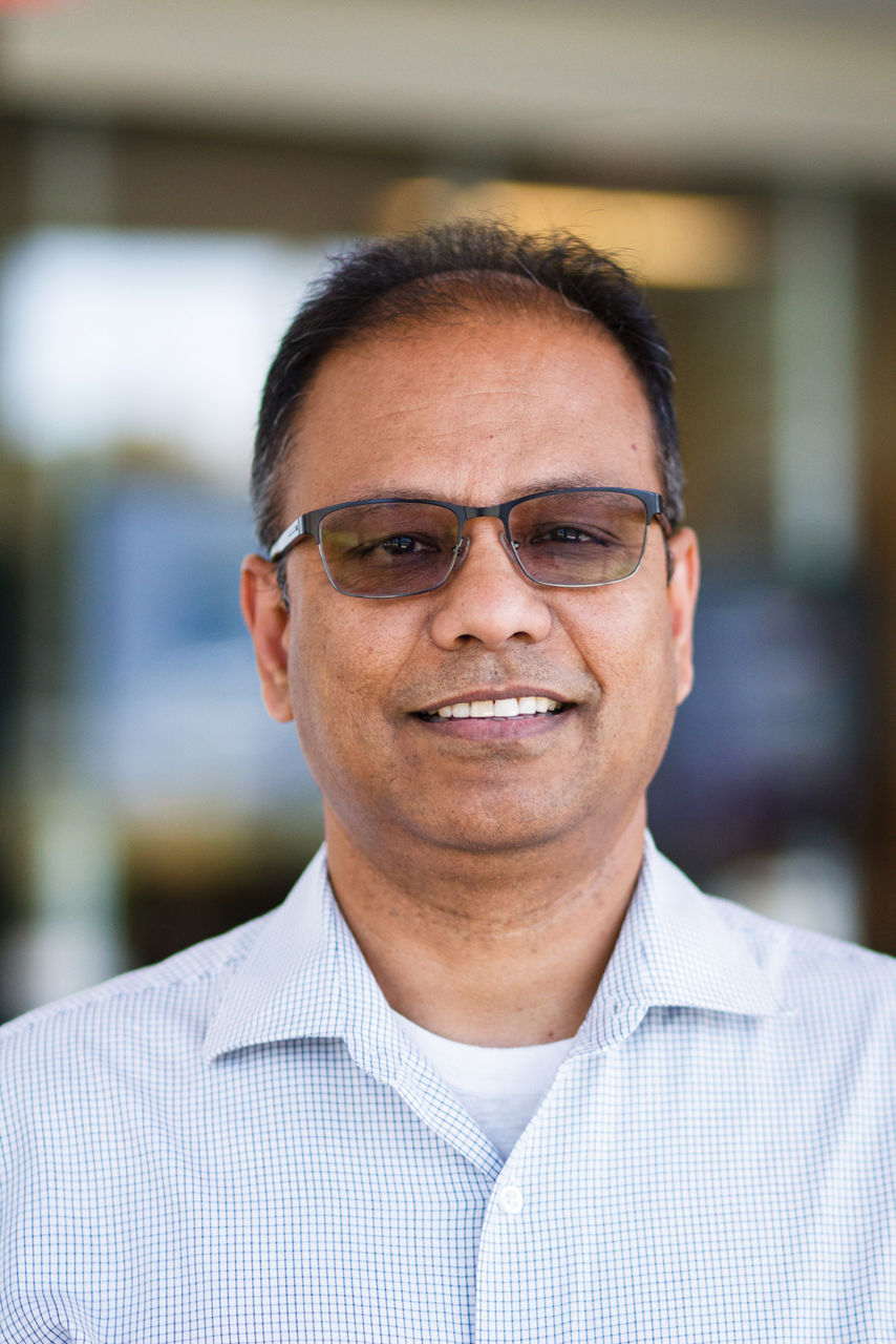A man wears a light blue checkered button-down shirt and glasses with transition lenses and smiles at the camera.