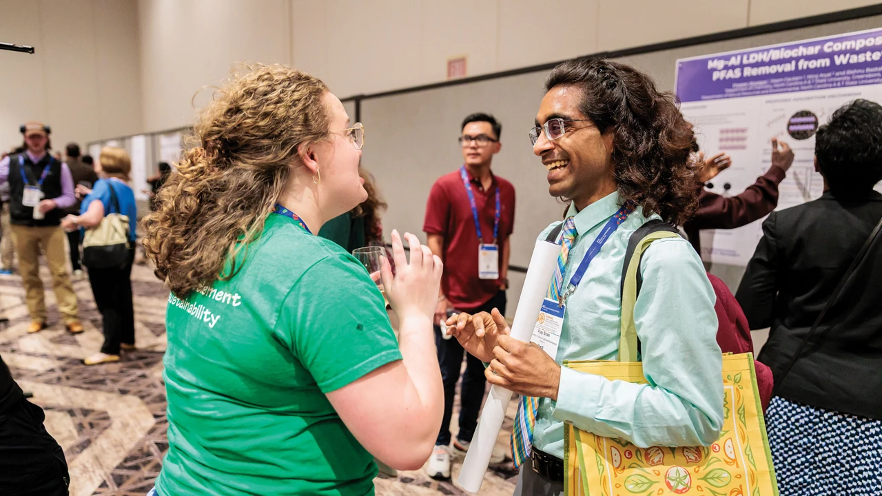 One person wearing a green shirt looks at another person wearing a light-blue collared shirt and holding a poster as both smile and laugh at each other in a room with other people in the background.