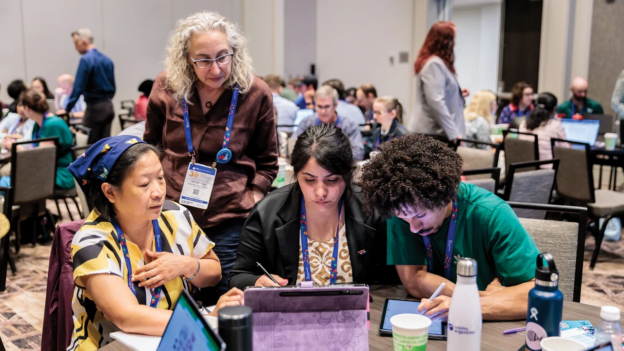 Three people sit at a table with open laptops and drinks and work on writing while another person stands and looks over their work, with additional tables and people in the background.