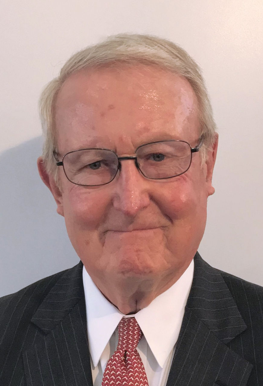 Ben wears a black pinstripe suit blazer, a tie, and white collared shirt, thin framed eyeglasses, and smiles at the camera.